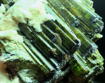 May include: A cluster of green and black tourmaline crystals with a white matrix. The crystals are long and slender, with a striated surface. The matrix is rough and textured.
