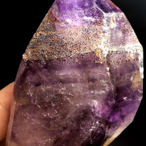 Pode incluir: Um grande cristal de ametista roxo com uma ponta. O cristal tem uma camada de inclusões douradas e marrons.