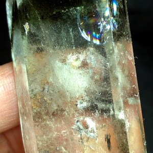 May include: A clear quartz crystal with a green inclusion and a rainbow iridescence. The crystal is pointed at the top and has a rough, natural base.