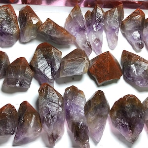 May include: A collection of amethyst crystals with a reddish-brown coating. The crystals are of various sizes and shapes, with some being pointed and others being more rounded. The crystals are arranged on a white surface.