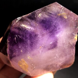 Pode incluir: Um grande cristal de ametista facetado com uma cor roxa profunda e uma tonalidade roxa mais clara na parte inferior. O cristal tem uma textura natural e áspera e é translúcido.