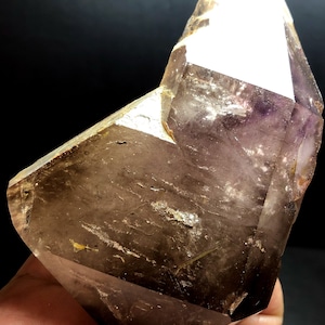 Pode incluir: Um grande cristal de quartzo transparente com um tom marrom esfumaçado e uma ponta de ametista roxa. O cristal tem uma forma facetada e é translúcido.