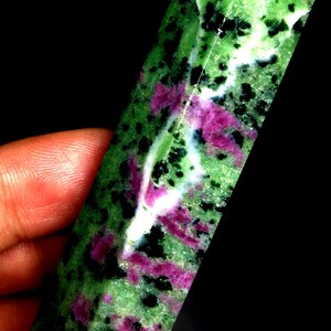 Op de afbeelding: Een groene, roze en witte edelsteen met zwarte vlekken in een lange, puntige vorm. De edelsteen is een natuurlijke kristal.