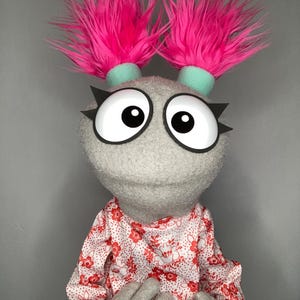 Może przedstawiać: Szara kukiełka z dużymi oczami z kreskówek, kolczastymi rzęsami i jasnoróżowymi, puszystymi kępkami włosów. Kukiełka ma na sobie koszulę z długim rękawem z białym tłem i czerwonym kwiatowym wzorem. Ręce kukiełki są szare.