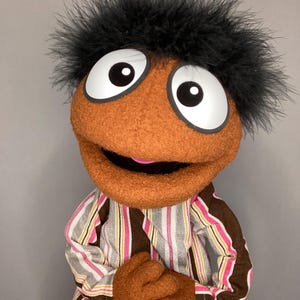 May include: A handmade puppet with a round, orange face, large white eyes, and a black feathery hair. The puppet wears a striped shirt with pink, brown, and white stripes and brown felt hands. The puppet's mouth is open, revealing a pink interior.