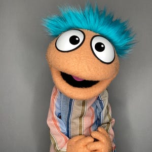 Peut inclure: Une marionnette faite à la main avec un visage couleur pêche, de grands yeux blancs avec des pupilles noires et des cheveux bleus vifs et hirsutes. La marionnette porte une chemise à rayures dans les tons de bleu, d'orange et de blanc. La bouche de la marionnette est ouverte, révélant une langue rose.