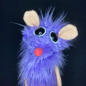 Może przedstawiać: Ręcznie robiona fioletowa kukiełka o puszystej fakturze, dużych czarnych oczach z białymi obrysami i czerwonym nosem. Kukiełka ma beżowe uszy i ramiona, a jej dłonie są złączone. Tło jest czarne.