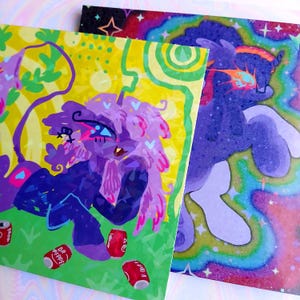 May include: Two vibrant art prints showcasing fantastical creatures. One print features a purple creature with pink hair and Dr. Pepper cans. The other print has a purple creature with a rainbow background and stars.