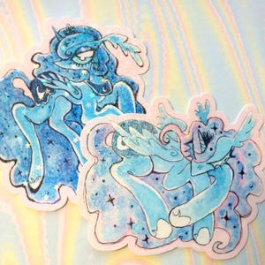 May include: Two stickers featuring a light blue cartoon pony with a flowing mane and tail, adorned with stars and glitter. The stickers have a white border and are set against a holographic background.