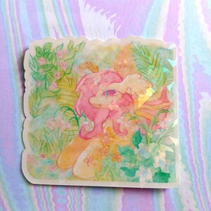 May include: A square-shaped sticker with a scalloped edge featuring a watercolour-style illustration of a pink-maned creature surrounded by lush green foliage, yellow flowers, and soft pastel hues with a subtle holographic finish throughout.