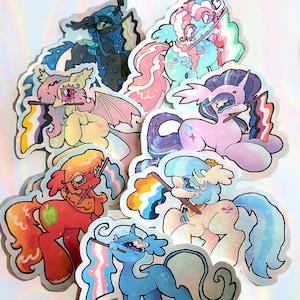 May include: A collection of holographic stickers featuring colorful cartoon ponies. Each pony holds a flag with a rainbow or pride flag design. The stickers have a glossy finish and are in various colors, including pink, blue, and yellow.