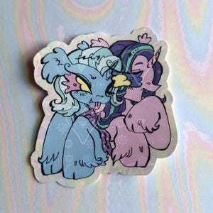 May include: A sticker featuring two cartoon characters, one blue and one purple, with a light blue background. The characters are hugging and have a friendly expression.