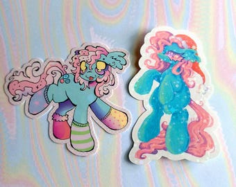 May include: Two pastel blue and pink cartoon pony stickers with glitter accents. One pony is wearing a Santa hat and the other is wearing socks.