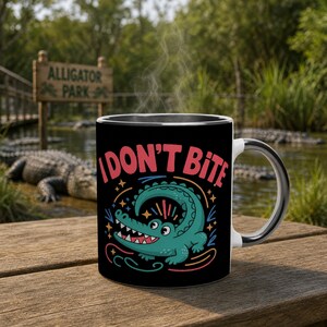 May include: Black ceramic mug with a white handle and rim, featuring a cartoon alligator design and the text "I DON'T BITE" in red. The mug is set on a wooden surface with an alligator park in the background.