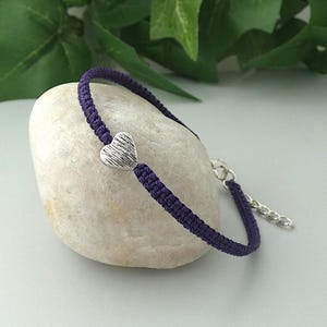 May include: A purple braided cord bracelet featuring a small, silver-toned heart-shaped charm with a textured surface. The bracelet includes a silver-toned metal lobster claw clasp and an adjustable extender chain for a secure fit around the wrist.