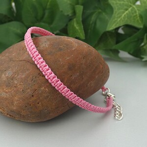 May include: A pink woven cord bracelet featuring a square knot pattern. The bracelet includes a silver-toned metal lobster claw clasp and an adjustable chain link extension, providing a secure and customisable fit for the wearer's wrist.