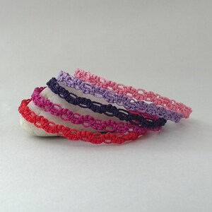 May include: A stack of five woven cord bracelets in red, magenta, dark blue, lavender, and light pink. Each bracelet features a repeating knotted pattern made from thin thread. The bracelets are arranged in a layered, overlapping formation.