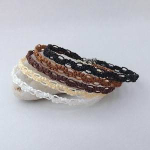 May include: A selection of macrame bracelets in shades of white, cream, brown, and black. The bracelets are arranged on a small, pale stone. Each bracelet has a woven pattern and a metal clasp. The bracelets are suitable for wrists of various sizes.