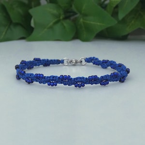 May include: A royal blue macrame bracelet featuring a series of small, matching blue beads woven into the cord. The bracelet is secured with a silver-toned metal lobster claw clasp and jump ring, creating a continuous, textured circular band.