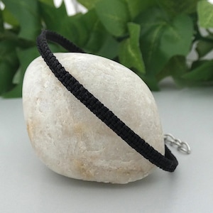 May include: A black cord bracelet featuring a woven macrame pattern and a silver-toned metal lobster claw clasp with an adjustable chain link extension. The bracelet is displayed in a circular shape, showcasing the uniform texture of the cord.