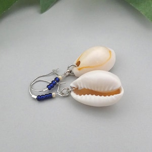 May include: A pair of seashell earrings. Each earring features a white and tan cowrie shell suspended from a silver hoop. Small blue beads and white beads are strung along the bottom of the hoop.