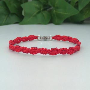 May include: A red macramé bracelet with a silver clasp. The bracelet is made of woven red threads, forming a repeating pattern. The bracelet is displayed on a white surface with green leaves in the background. A piece of jewellery.
