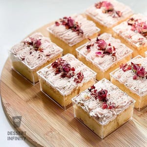 May include: Nine square cakes in clear plastic containers, arranged on a wooden board. Each cake has a light-colored base, topped with swirled pink frosting, dried rose petals, and herbs. The cakes are labeled "Dessert Infinity".