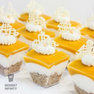 Puede incluir: Primer plano de varios postres individuales en vasos cuadrados transparentes. Cada postre tiene una base de migas, una capa blanca y una parte superior de color naranja brillante, cubierta con crema batida blanca y decoraciones de chocolate blanco. El texto "DESSERT INFINITY" es visible.