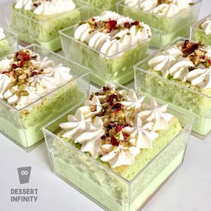 Może przedstawiać: Indywidualne desery w przezroczystych kwadratowych pojemnikach. Każdy deser ma jasnozieloną podstawę, zwieńczoną białą bitą śmietaną, posiekanymi orzechami i suszonymi owocami. Logo "Dessert Infinity" jest widoczne w lewym dolnym rogu.