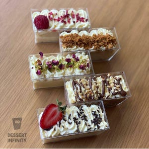 以下が含まれることがあります： 透明なプラスチック容器に入った長方形のデザート5つの俯瞰図。各デザートは白いクリームトッピングで、ラズベリー、ナッツ、チョコレートドリズルなどの様々な飾り付けがされています。「Dessert Infinity」の文字が見えます。