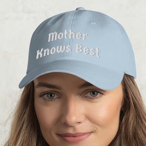 May include: Light blue baseball cap with the embroidered text "Mother Knows Best" in white. The cap has a curved brim and a classic six-panel design. The hat is made of a soft, breathable fabric.