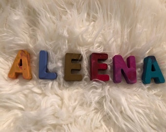 Puede incluir: El nombre "ALEENA" deletreado en bloques de letras de cera multicolores. Los bloques están dispuestos en línea sobre una superficie blanca y esponjosa.