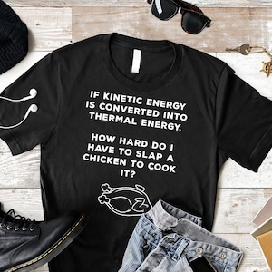 Op de afbeelding: Zwart T-shirt met witte tekst die luidt "ALS KINETISCHE ENERGIE WORDT OMGEZET NAAR THERMISCHE ENERGIE, HOE HARD MOET IK EEN KIP SLAN OM HEM TE KOKEN?" met een cartoon-kipillustratie.