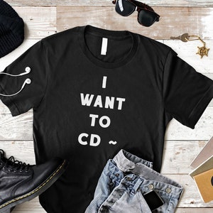 May include: Black t-shirt with white text that reads "I WANT TO CD ~". The shirt is laid out on a white wood surface with a beanie, sunglasses, jeans, and a pair of black boots.