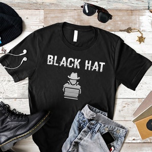 Puede incluir: Camiseta negra con un gráfico blanco de una persona con un sombrero sentada frente a una computadora con el texto "BLACK HAT" sobre el gráfico.