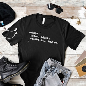 May include: Black t-shirt with white text reading ".ninja { color: black; visibility: hidden; }". The shirt is styled with blue jeans, a black beanie, black boots, and sunglasses. White earbuds are placed on the shirt.