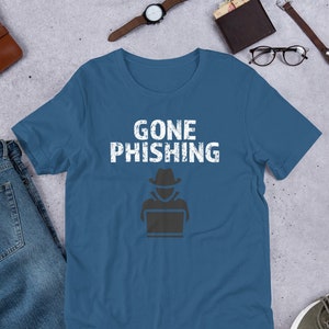 Könnte beinhalten: Ein blaues T-Shirt mit dem Text "GONE PHISHING" in Weiß mit einem Used-Look. Unter dem Text befindet sich eine schwarze Silhouette einer Person mit Hut, die einen Laptop benutzt.