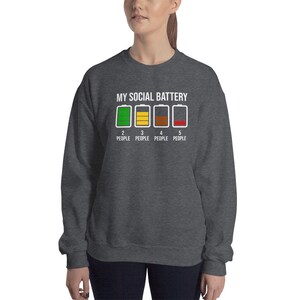May include: A dark grey sweatshirt with the text "MY SOCIAL BATTERY" and a graphic of four battery icons with different levels of charge. The text below the icons reads "2 PEOPLE, 3 PEOPLE, 4 PEOPLE, 5 PEOPLE".