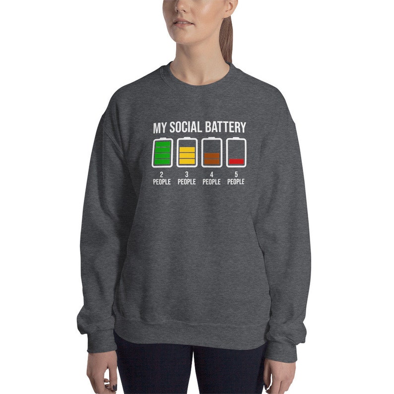 May include: A dark grey sweatshirt with the text "MY SOCIAL BATTERY" and a graphic of four battery icons with different levels of charge. The text below the icons reads "2 PEOPLE, 3 PEOPLE, 4 PEOPLE, 5 PEOPLE".