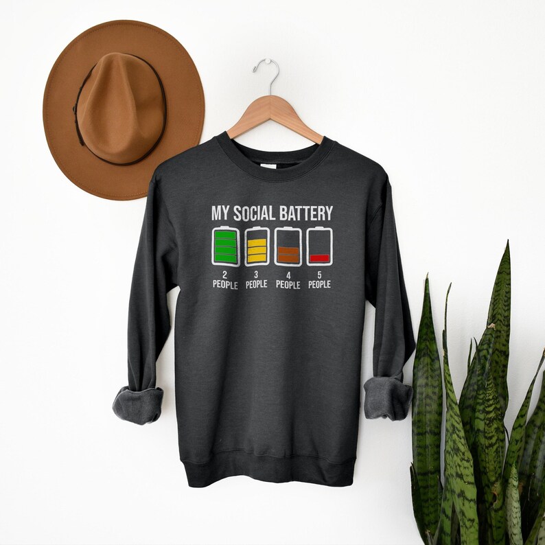 May include: A dark grey sweatshirt with the text "My Social Battery" and a graphic of four battery icons with different levels of charge. The text "2 People", "3 People", "4 People", and "5 People" is below the battery icons.
