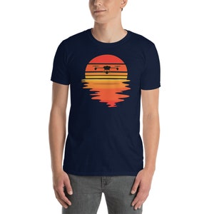 Puede incluir: Camiseta azul marino con un dise&ntilde;o de atardecer y dron. El dise&ntilde;o presenta una silueta de dron negra sobre un vibrante degradado naranja y amarillo, con un reflejo debajo. La camiseta tiene cuello redondo y mangas cortas.