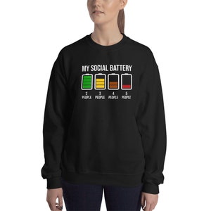 May include: Black sweatshirt with a white graphic of a battery meter labeled "My Social Battery" with different colored bars representing different levels of social energy. The text below the battery meter reads "2 People, 3 People, 4 People, 5 People".