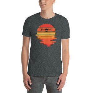 Puede incluir: Camiseta gris oscuro con una silueta de dron sobre un dise&ntilde;o de puesta de sol. La puesta de sol est&aacute; representada en tonos naranja y amarillo, con un efecto de reflejo debajo. La camiseta es de un material suave.