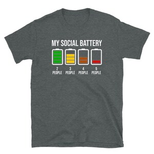 May include: A dark gray t-shirt with the text "MY SOCIAL BATTERY" and a graphic of five battery icons. The battery icons are labeled with numbers 2 through 5 and the text "PEOPLE". The first battery icon is green, the second is yellow, the third is brown, and the fourth is red.