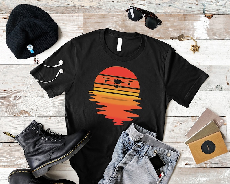 Puede incluir: Camiseta negra con un dise&ntilde;o de dron sobre un atardecer naranja y amarillo. La imagen incluye un gorro negro, botas negras, gafas de sol y vaqueros. El dise&ntilde;o de la camiseta es un gr&aacute;fico.
