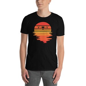 Puede incluir: Camiseta negra con un dise&ntilde;o gr&aacute;fico de un dron sobre un dise&ntilde;o de puesta de sol. El dron est&aacute; representado en silueta, con el sol en tonos naranja y amarillo. El dise&ntilde;o incluye un reflejo de la puesta de sol debajo.