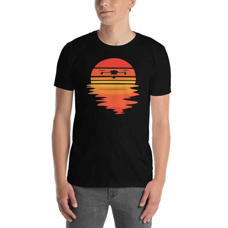 Puede incluir: Camiseta negra con un dise&ntilde;o gr&aacute;fico de un dron sobre un dise&ntilde;o de puesta de sol. El dron est&aacute; representado en silueta, con el sol en tonos naranja y amarillo. El dise&ntilde;o incluye un reflejo de la puesta de sol debajo.