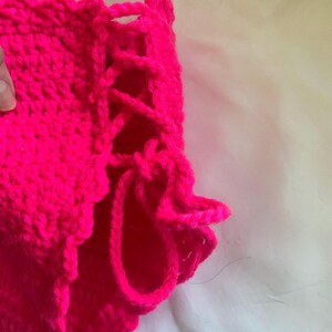 Puede incluir: Una prenda de ganchillo en color rosa intenso que presenta un detalle de cordones laterales con lazos de hilo grueso. La pieza est&aacute; confeccionada con un patr&oacute;n de punto denso y texturizado, e incluye un borde festoneado en el dobladillo inferior.