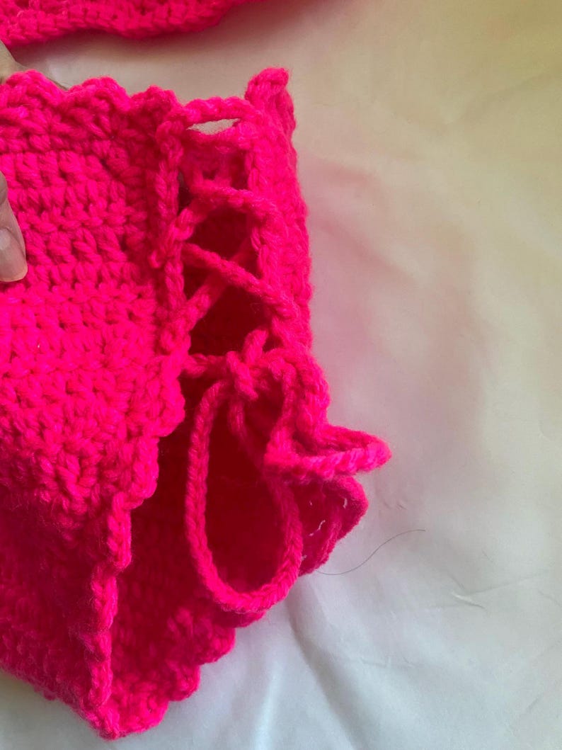Puede incluir: Una prenda de ganchillo en color rosa intenso que presenta un detalle de cordones laterales con lazos de hilo grueso. La pieza est&aacute; confeccionada con un patr&oacute;n de punto denso y texturizado, e incluye un borde festoneado en el dobladillo inferior.