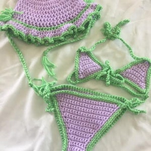 Puede incluir: Un conjunto de ganchillo compuesto por un sombrero de pescador color lavanda con ribete de volantes verde claro, un top de bikini triangular lavanda con lazos verde claro y una braguita de bikini lavanda a juego con ribete y lazos laterales.